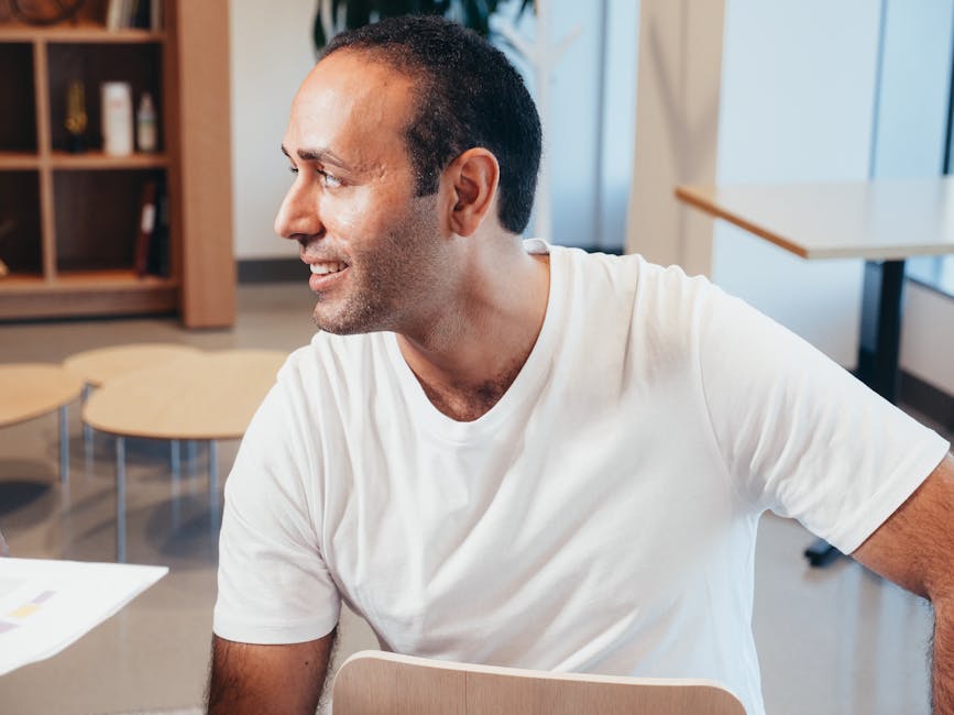 A confident man smiling in a modern office space, wearing a white t-shirt.